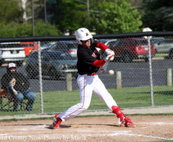 Baseball player in black and red swings at a pitch on a dirt field with a chain-link fence in the background.