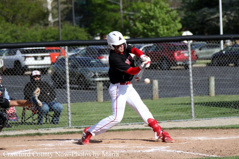 Baseball player in black and red swings at a pitch on a dirt field with a chain-link fence in the background.