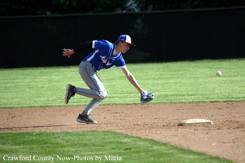 Baseball infielder in a blue uniform lunges to field a ground ball near first base on a sunny field.
