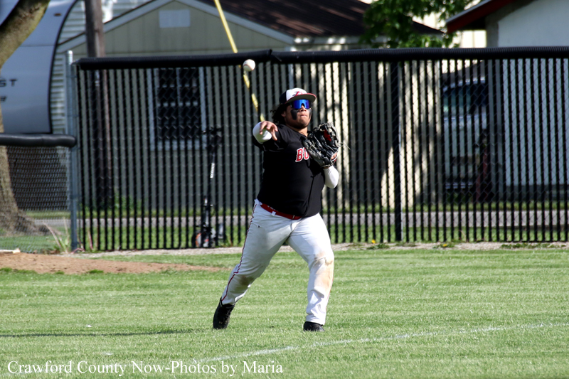 Baseball fielder in a black uniform and white pants throws a ball on a grassy field, with a chain-link fence and buildings in the background.