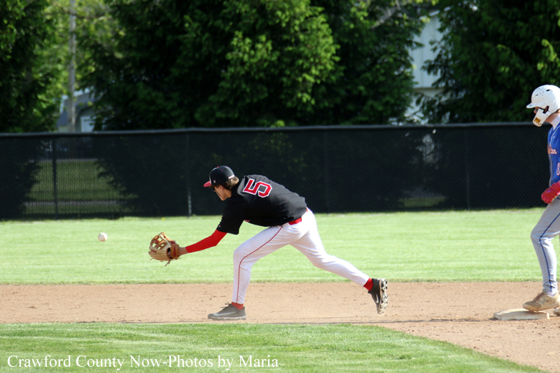 Baseball infielder in a black jersey with red sleeves and number 5 lunges to field a ground ball on the infield as a runner watches from the right side of the frame.