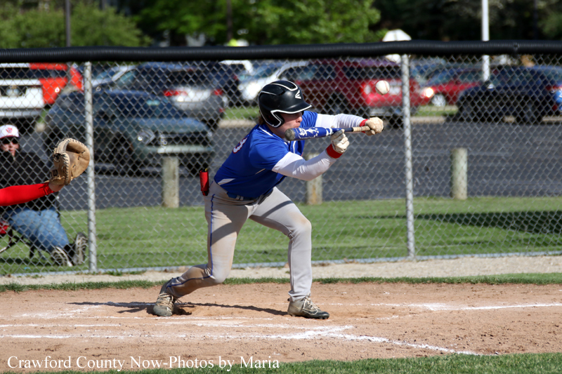 Baseball player in blue swings at a pitch at home plate, catcher crouched behind him near a chain-link fence.