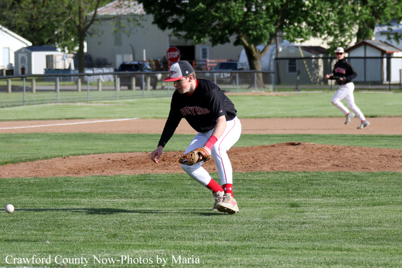 Baseball pitcher in black jersey crouches to field a ground ball on the grass and dirt infield; fielder in background.
