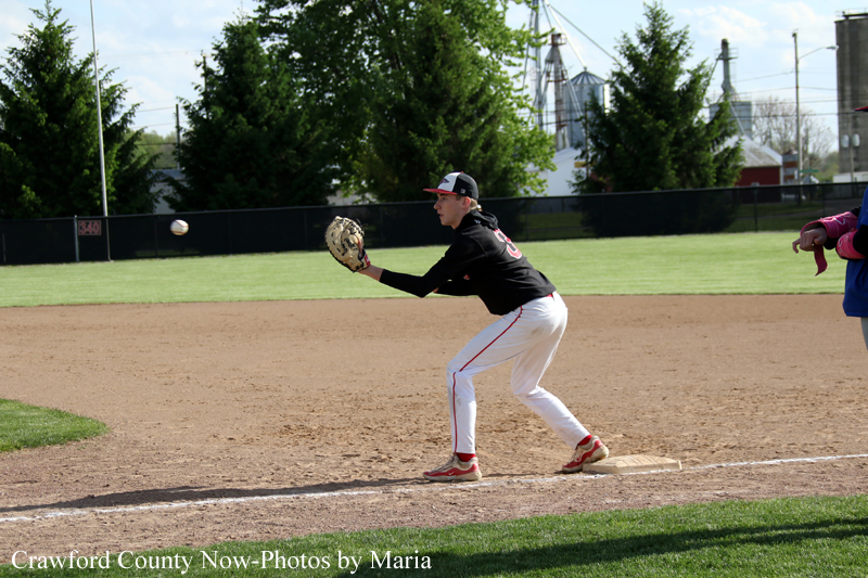 Baseball player in a black jersey and white pants fields a ground ball near first base, glove ready, as an umpire watches in the background.