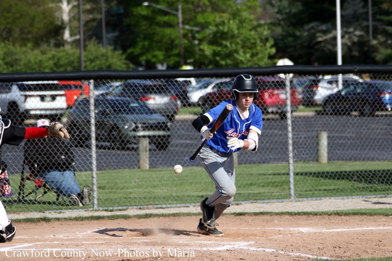 Young baseball player in a blue uniform and helmet runs toward first base as a pitch nears, with a catcher and a chain-link fence in the background.