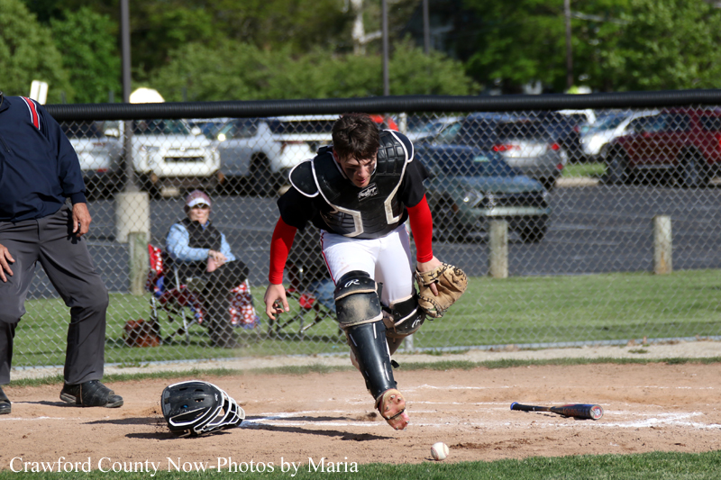Youth baseball catcher lunges toward home plate as a ball rolls away, helmet and bat lying on the dirt nearby, spectators behind a chain-link fence in a sunny field.