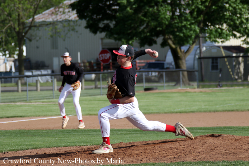 Baseball pitcher in black and red uniform delivering a pitch on the mound, with a fielder in the background.