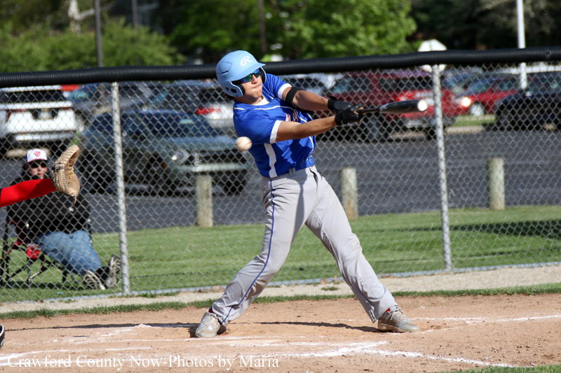 Baseball batter in a blue uniform swings at a pitched ball at home plate, wearing a helmet and batting gloves as a catcher sits behind the fence in the background.