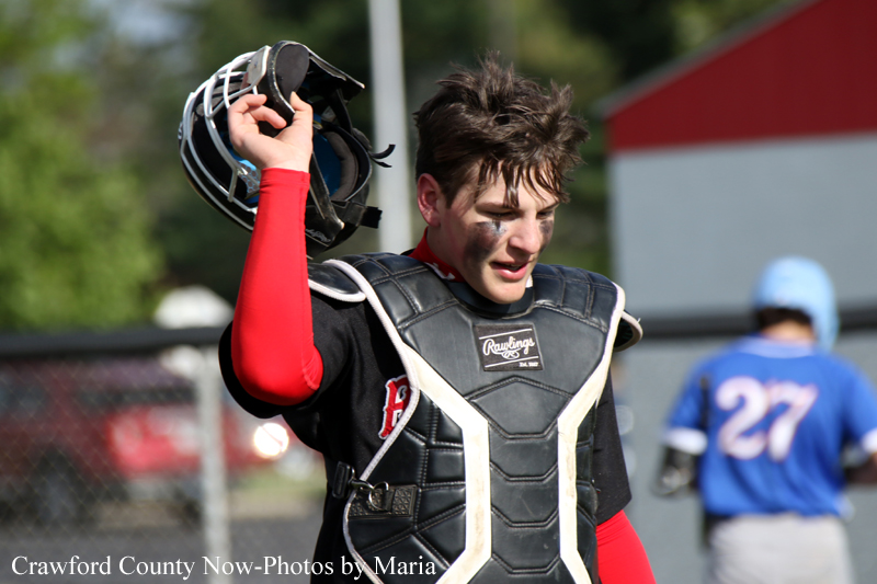 Baseball catcher in protective gear lifts his helmet after a play on the field, red undershirt visible.