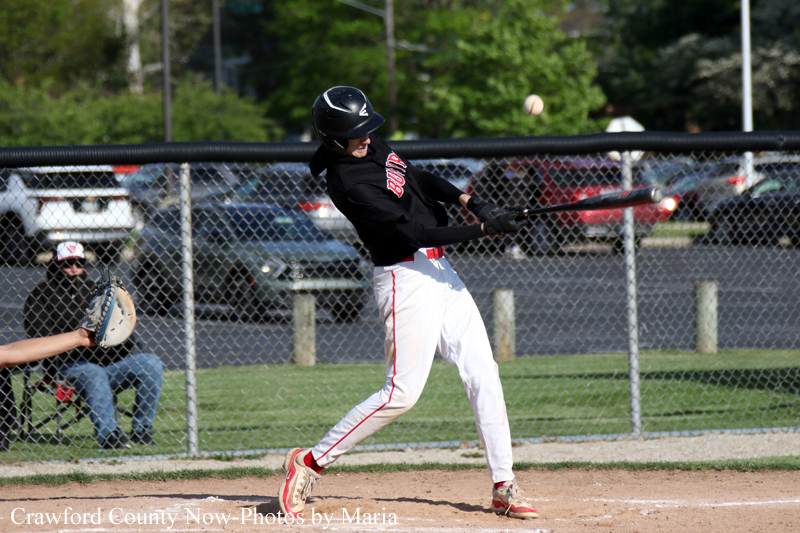 Baseball batter in black helmet swings at a pitch during a game on a dirt infield with a chain-link fence in the background.