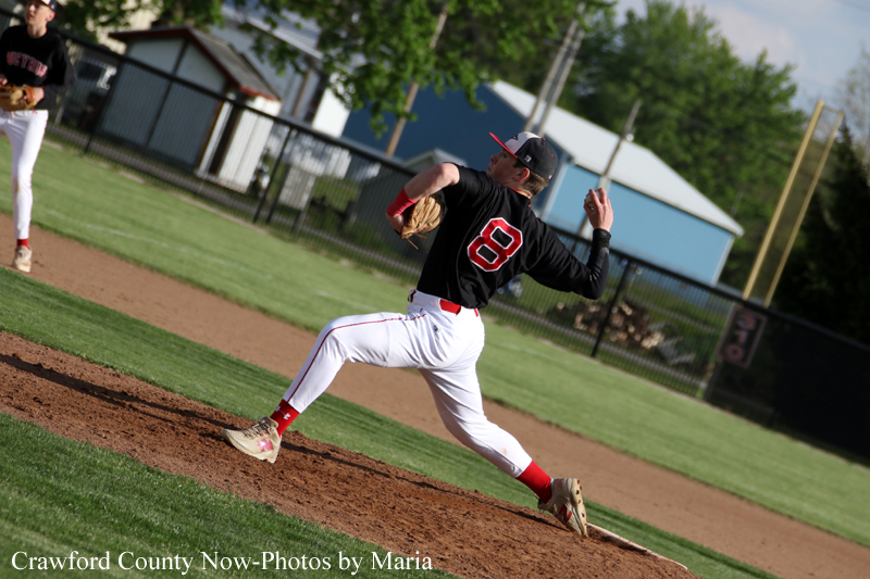 Baseball pitcher in a black jersey with the number 8 winds up on the mound, throwing a pitch with a teammate visible in the background.
