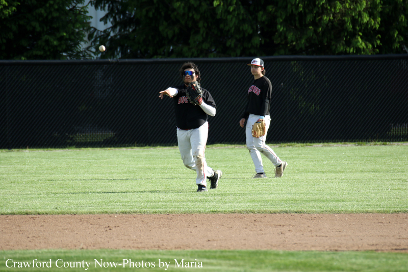 Two baseball players on a green field, one pitcher throwing a ball while a teammate watches near a dark outfield fence.