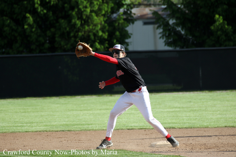 Baseball pitcher mid-pitch on a dirt infield, wearing a black jersey with red sleeves and white pants, gloves the ball as he throws on a sunny day.