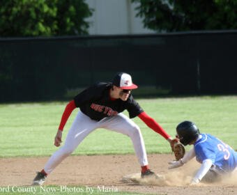 Baseball field scene: a fielder in black and red leans to tag a sliding baserunner in blue as dust rises at the base edge.