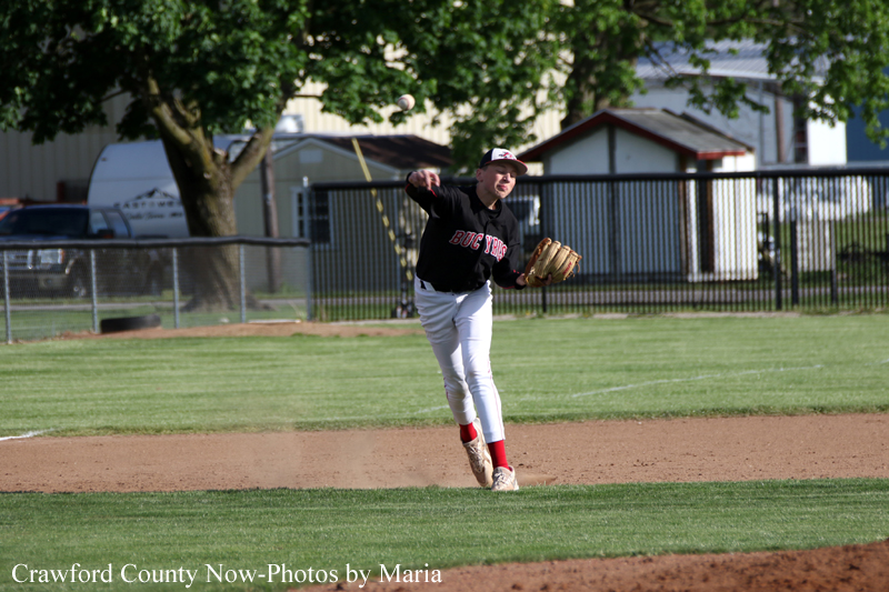 Baseball pitcher in a black jersey and white pants mid-throw on the infield, ball in the air with a glove ready at his side.