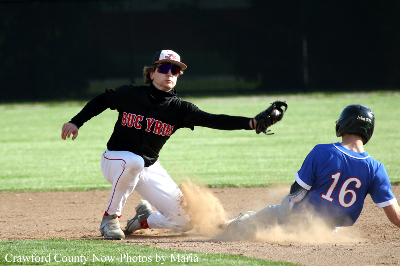 Baseball scene: fielder in black raises glove toward a sliding baserunner in blue #16 as dirt flies around them on a dusty infield.
