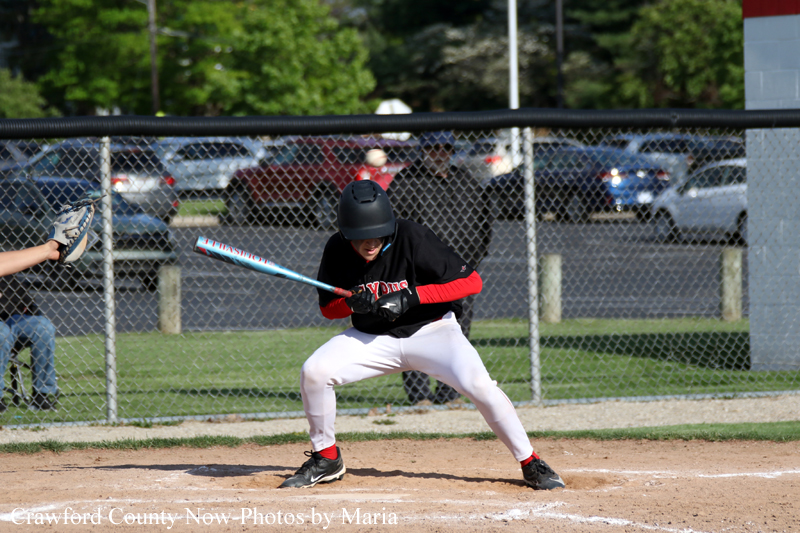 Baseball batter in a black jersey and white pants crouches to swing, helmet on, on a dirt infield with a chain-link fence and spectators behind.