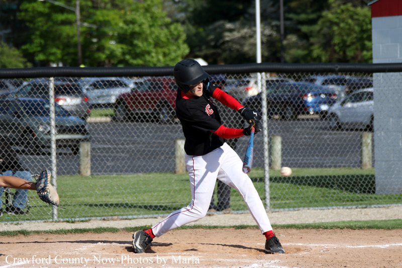 Baseball batter in black and red uniform swings at a pitched ball at home plate, with a catcher’s leg visible to the left and a chain-link fence in the background.