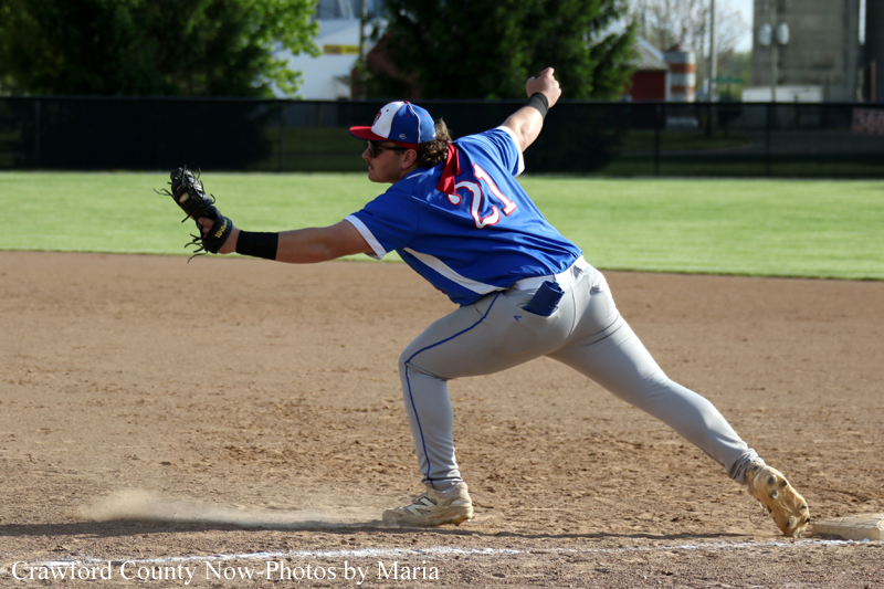 Baseball pitcher in blue jersey delivering a pitch from the infield mound with dust rising near home plate.