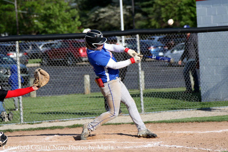 Baseball batter in a blue jersey and helmet swings at a pitch on a dirt field with a catcher nearby.