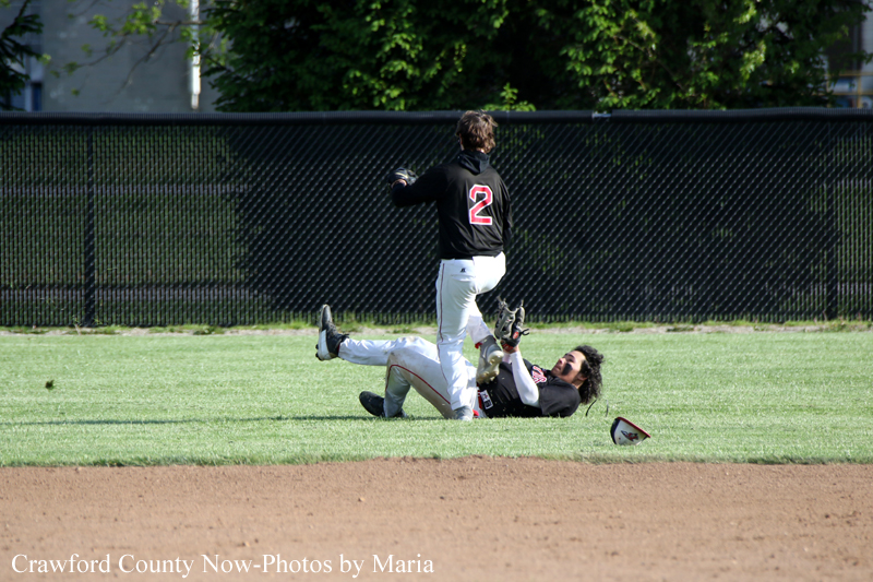Baseball field scene: one player with number 2 on a black jersey stands over a teammate who has slid and is lying on the grass, glove raised, with a helmet and glove nearby, a chain-link fence and trees in the background.