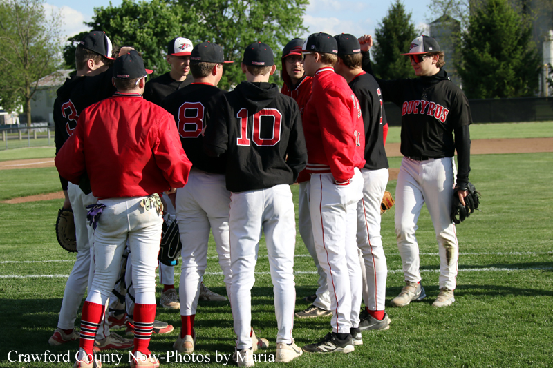 Baseball players in red and black uniforms form a huddle on the field on a sunny day