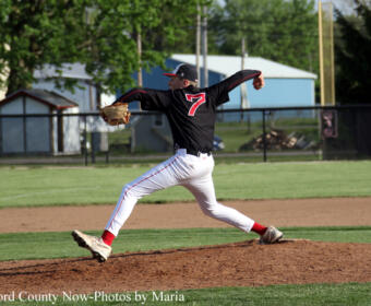 Baseball pitcher in a black jersey with number 7 delivers a pitch from the mound on a sunny field.
