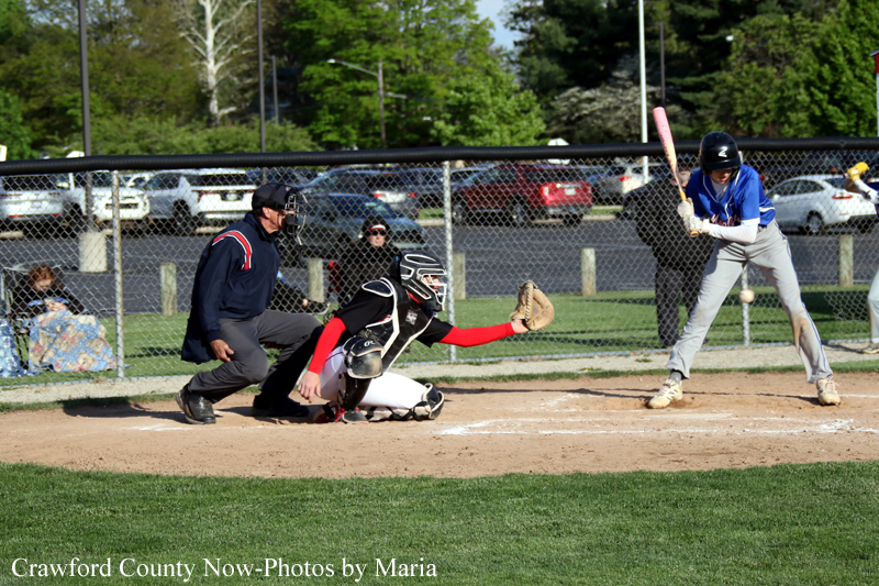 Blue-clad batter at home plate prepares to swing as the catcher in black/red gear and the umpire observe a softball play.