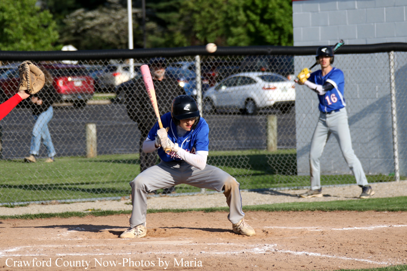 Youth baseball player in blue at home plate, bat raised and ready to swing as the pitch comes in. Behind her, a teammate in blue watches from the field fence.