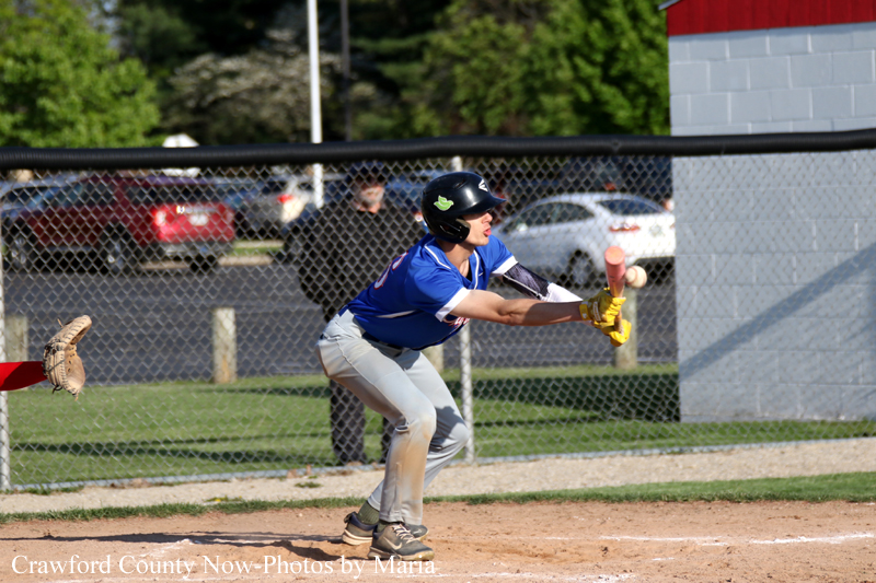 Batter in a blue uniform and helmet swings at a pitch on a dirt baseball field, with a catcher’s glove visible to the left and a chain-link fence in the background.