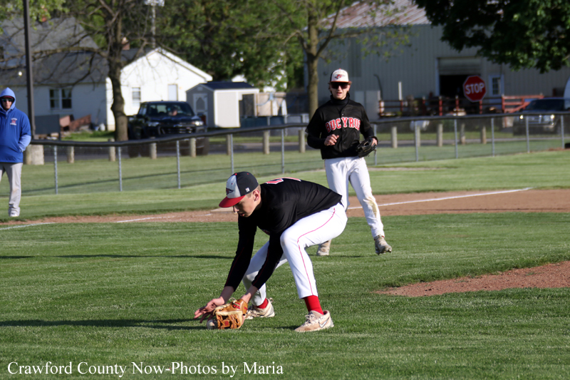 Infielder in a black jersey bends to field a ground ball with his glove close to the dirt on a sunny baseball field; a teammate watches in the background.