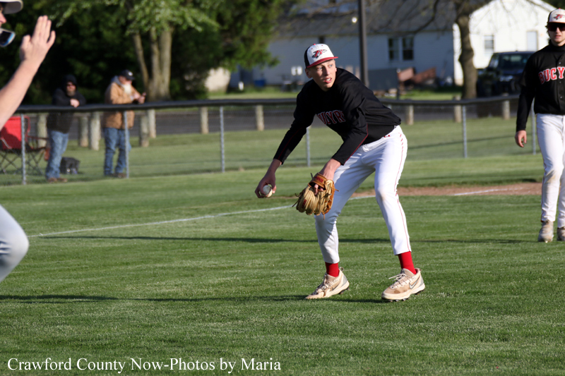 Baseball fielder in a black jersey and white pants crouches to field a ground ball, glove ready as spectators and a fence line the outfield in the background.