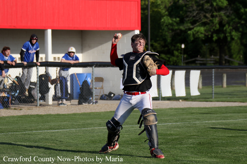 Baseball pitcher in black chest protector and white pants throwing a pitch on a sunny field, with teammates and a dugout in the background.