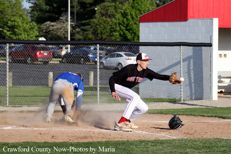 Two youth baseball players on a dirt infield: a fielder in black reaches for the ball while a blue-uniformed runner slides into base, kicking up dust as a helmet lies nearby.
