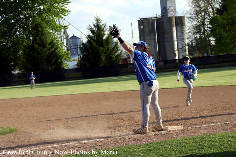 Baseball player in blue on first base with glove raised, another fielder running in the background on a sunny infield.
