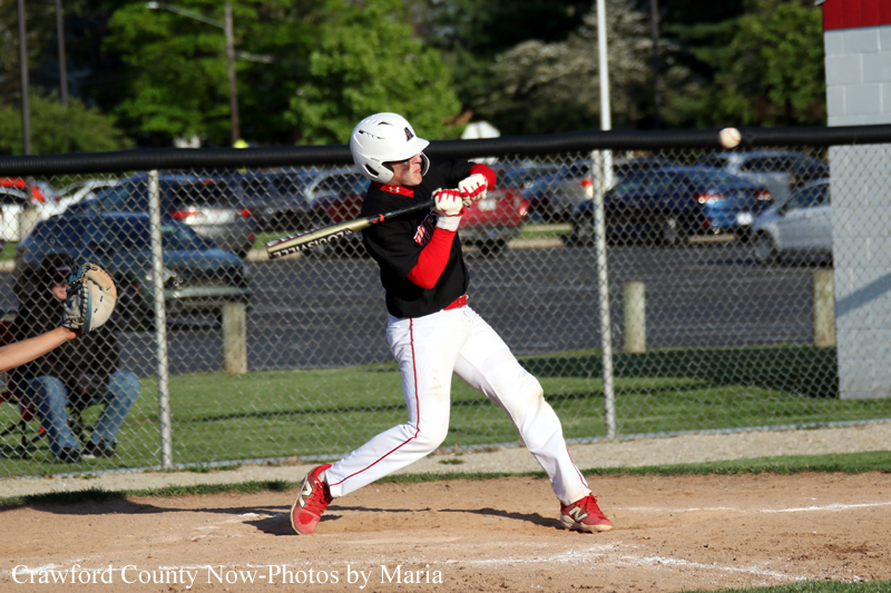 Baseball player in black and red uniform swings at a pitch during a game, with catcher behind him near the plate.