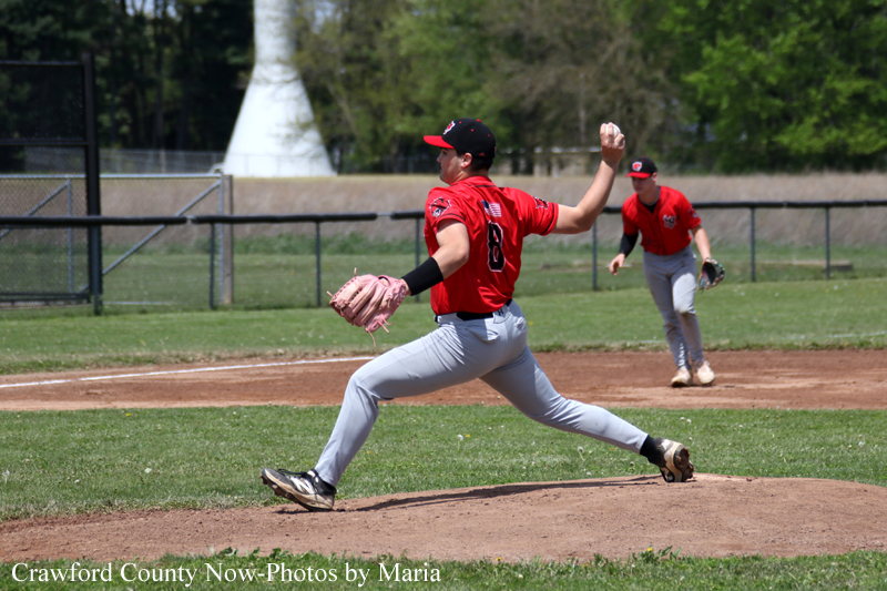 Baseball pitcher in red uniform winds up and throws from the mound as a fielder watches in the infield on a sunny field.