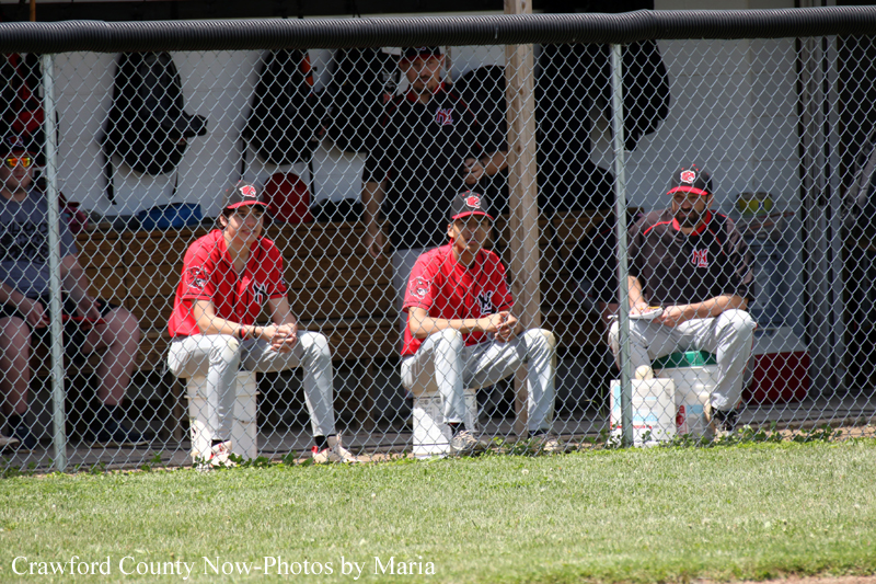 Baseball players in red jerseys sit on the dugout bench behind a chain-link fence during a game.