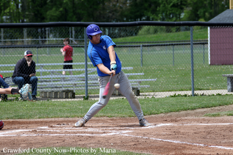 Baseball player in a blue helmet and shirt swings at a pitched ball on a dirt batter's box with a chain-link fence in the background.