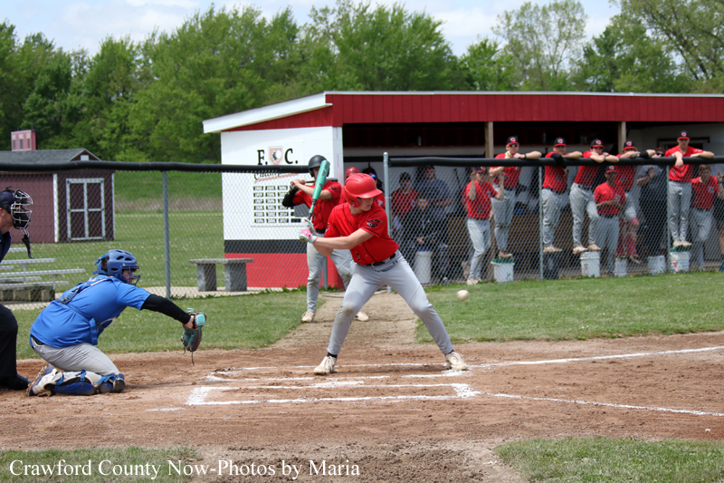 Red-jersey batter swings at a pitch at home plate, with a blue-uniformed catcher crouched behind him as teammates watch from the dugout.