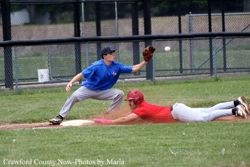 Baseball infield action: a blue-uniformed fielder lunges toward the base while a red-uniformed runner slides headfirst into the bag, dirt flying.