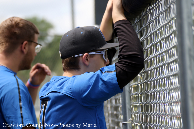 Two baseball players in blue uniforms lean on a chain-link fence, peering through toward the action outside the field.