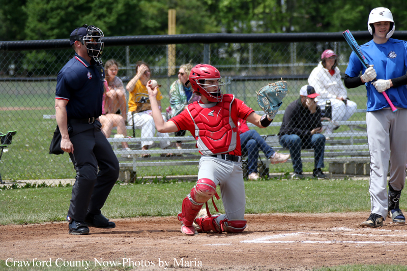 Youth baseball catcher in red gear slides to home plate as the umpire watches; batter in blue stands ready nearby with a bat, and spectators sit behind the chain-link fence.