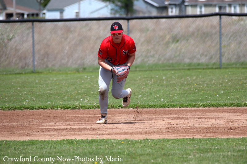 Baseball player in a red jersey and white pants fields a ground ball on the infield dirt with a chain-link fence behind him.