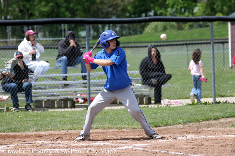 Young baseball batter in blue swings at a pitch on a dirt infield, with fans watching behind a chain-link fence.