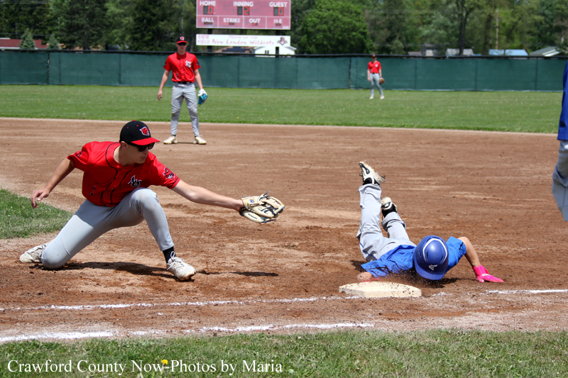 Fielder in red dives to tag a sliding baserunner in blue as they reach a base during a baseball game, with players and scoreboard in the background.