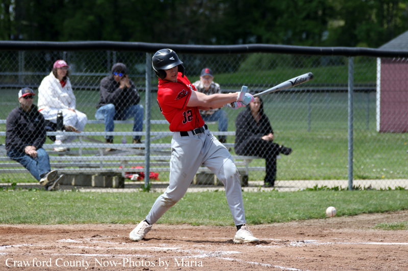 Baseball batter in a red uniform swings at a pitch on a dirt field, with spectators behind a chain-link fence.