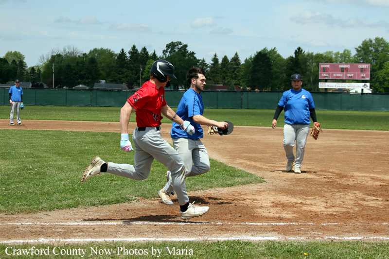 Two runners sprint toward first base as a fielder in blue looks on on a sunny baseball field.