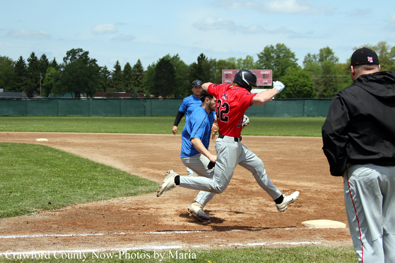Red-jersey baserunner #22 leaps toward a base as a blue-jersey fielder runs behind him on the dirt infield with an umpire watching from the right.