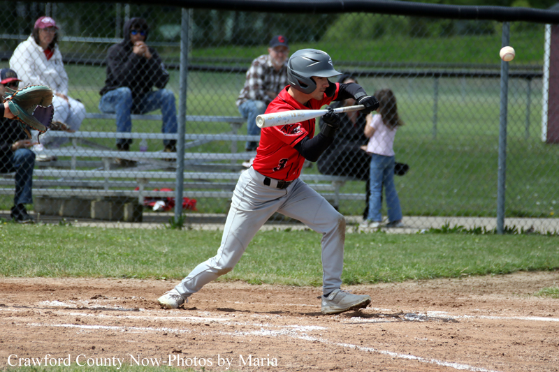 A young baseball player in a red jersey swings a bat at a pitched ball on a dirt batter's box with a chain-link fence and spectators in the background